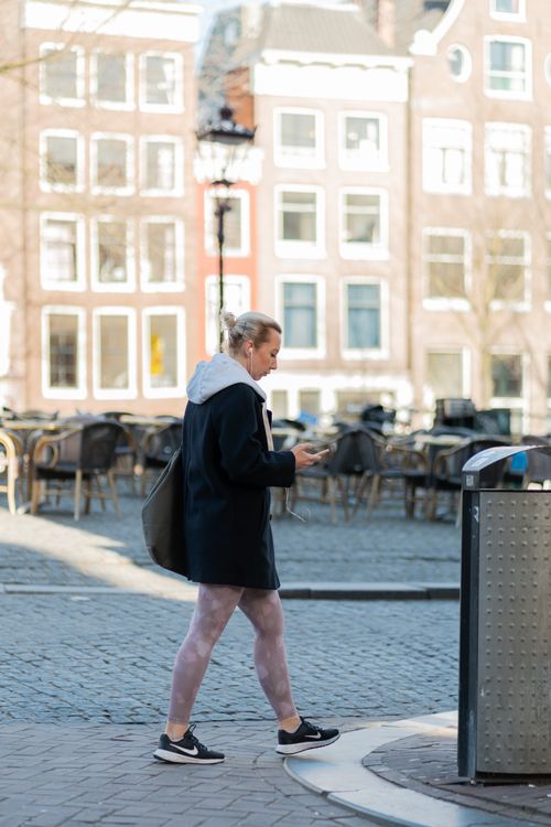 Free A woman walks through an urban square while using her smartphone, surrounded by cafe tables and classic architecture. Stock Photo
