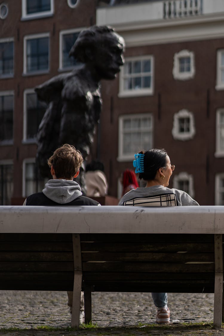 Boy And A Woman Sitting On A City Bench With A Statue In The Background