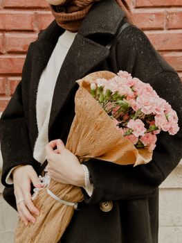 A woman in a dark coat holds a pink carnation bouquet against a red brick wall, creating a warm and stylish scene.