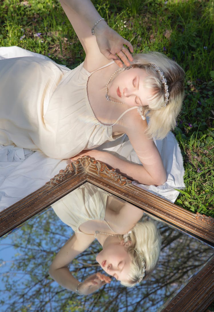 Young Woman In A White Dress Lying On The Grass With A Mirror 