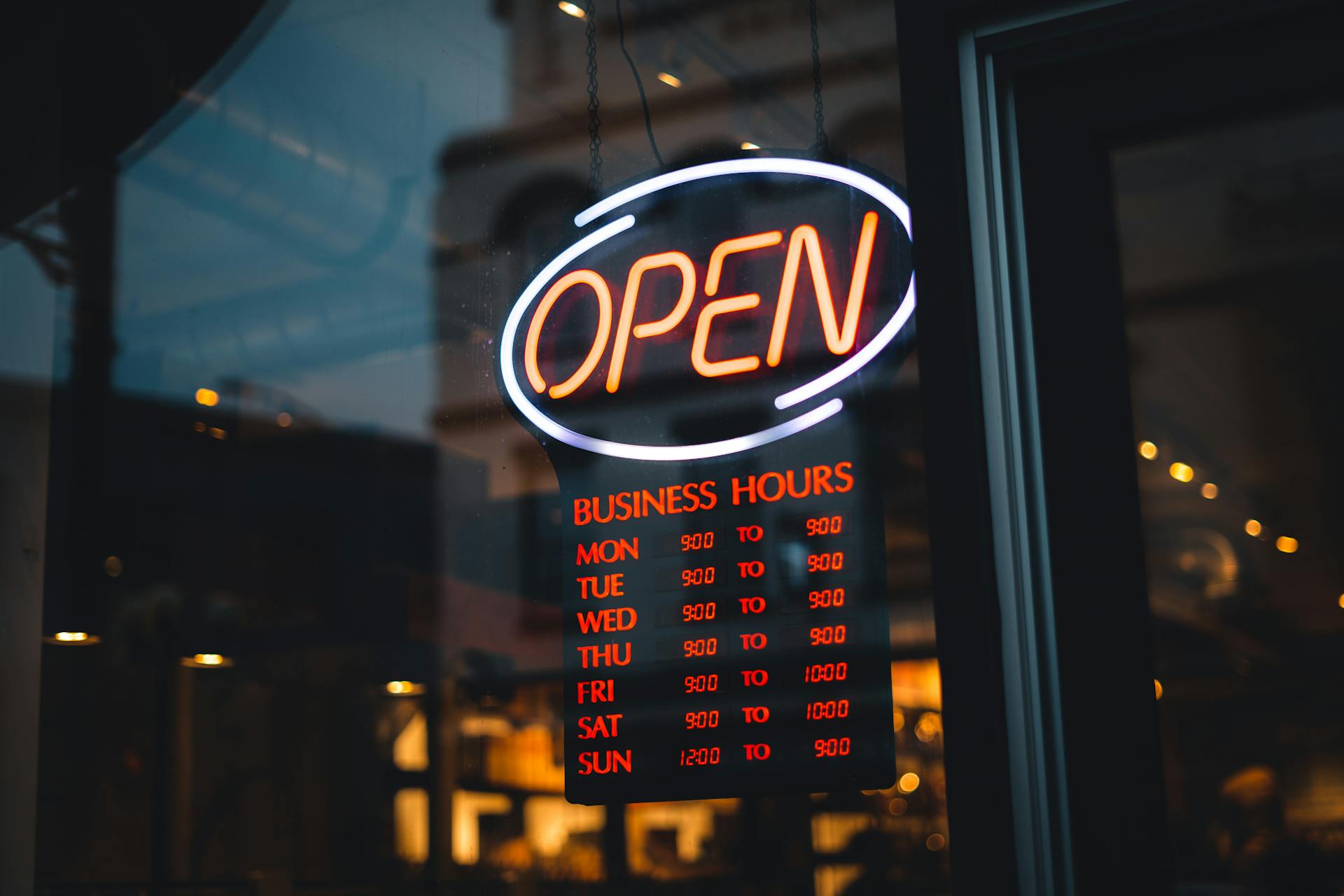 A glowing neon 'OPEN' sign with detailed business hours in a storefront window at night.