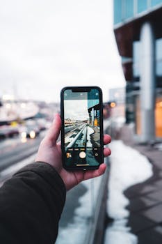 Hand holding smartphone photographing urban winter scene with buildings.