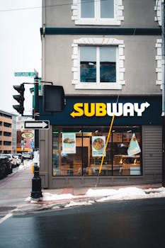 Exterior view of a Subway restaurant on a city street corner with snow and traffic lights.