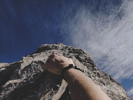 A climber's hand gripping a rocky cliff under a clear blue sky, embodying adventure.