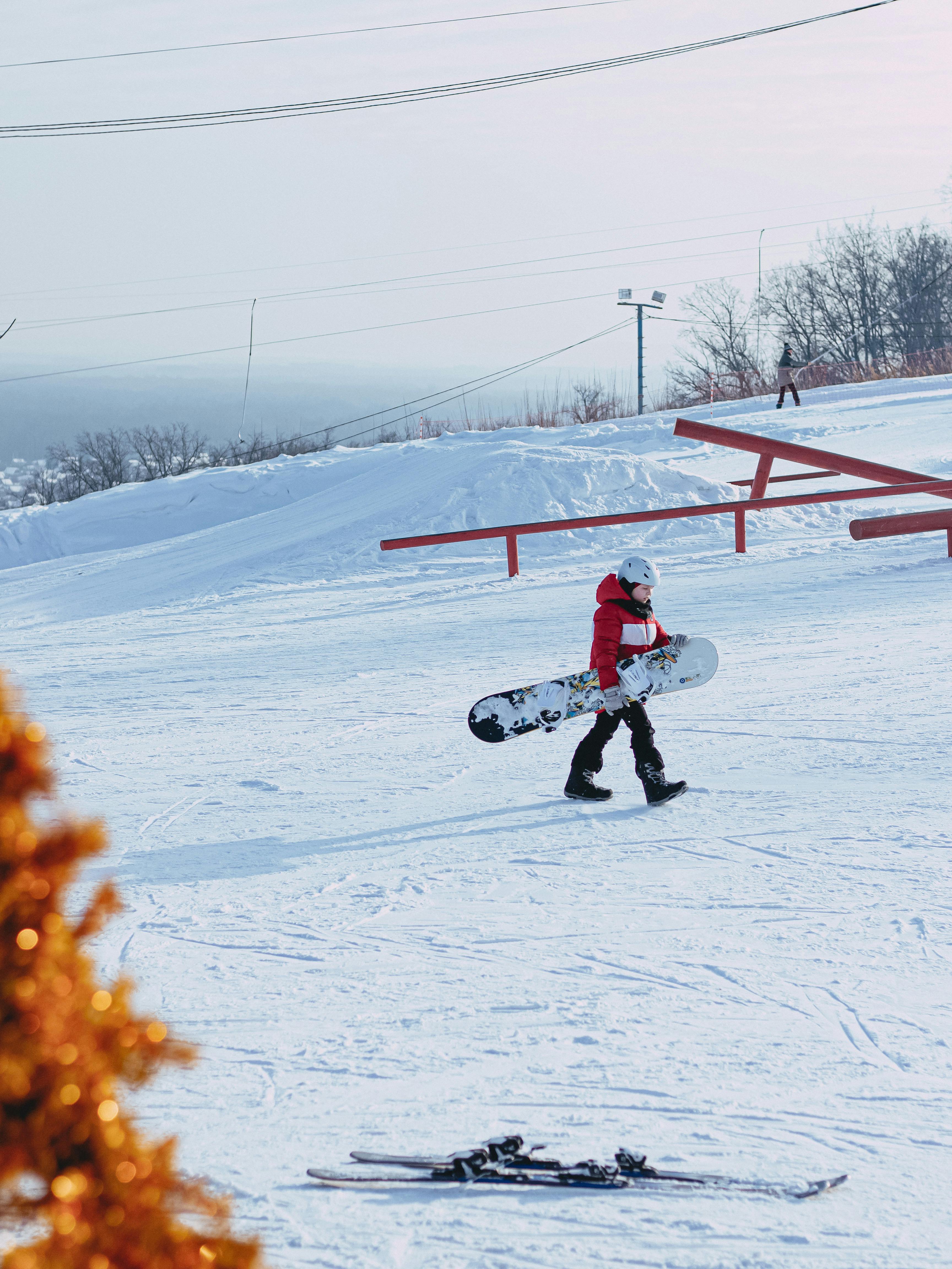 Prescription Goggle Inserts - A snowboarder in a red jacket walks uphill on a snowy slope, carrying a snowboard, during a winter day.