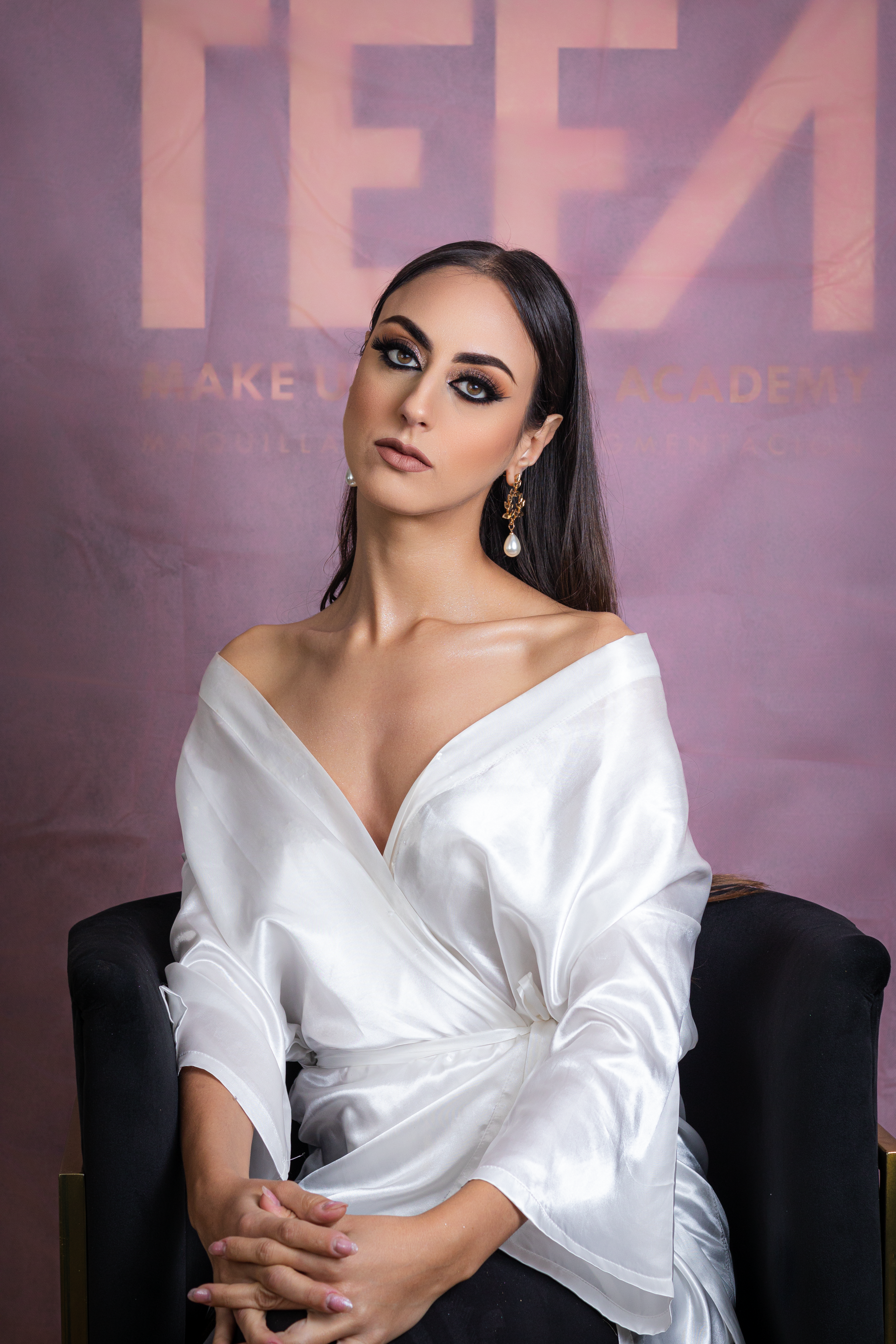 A glamorous woman posing indoors in a white satin dress, seated against a backdrop.
