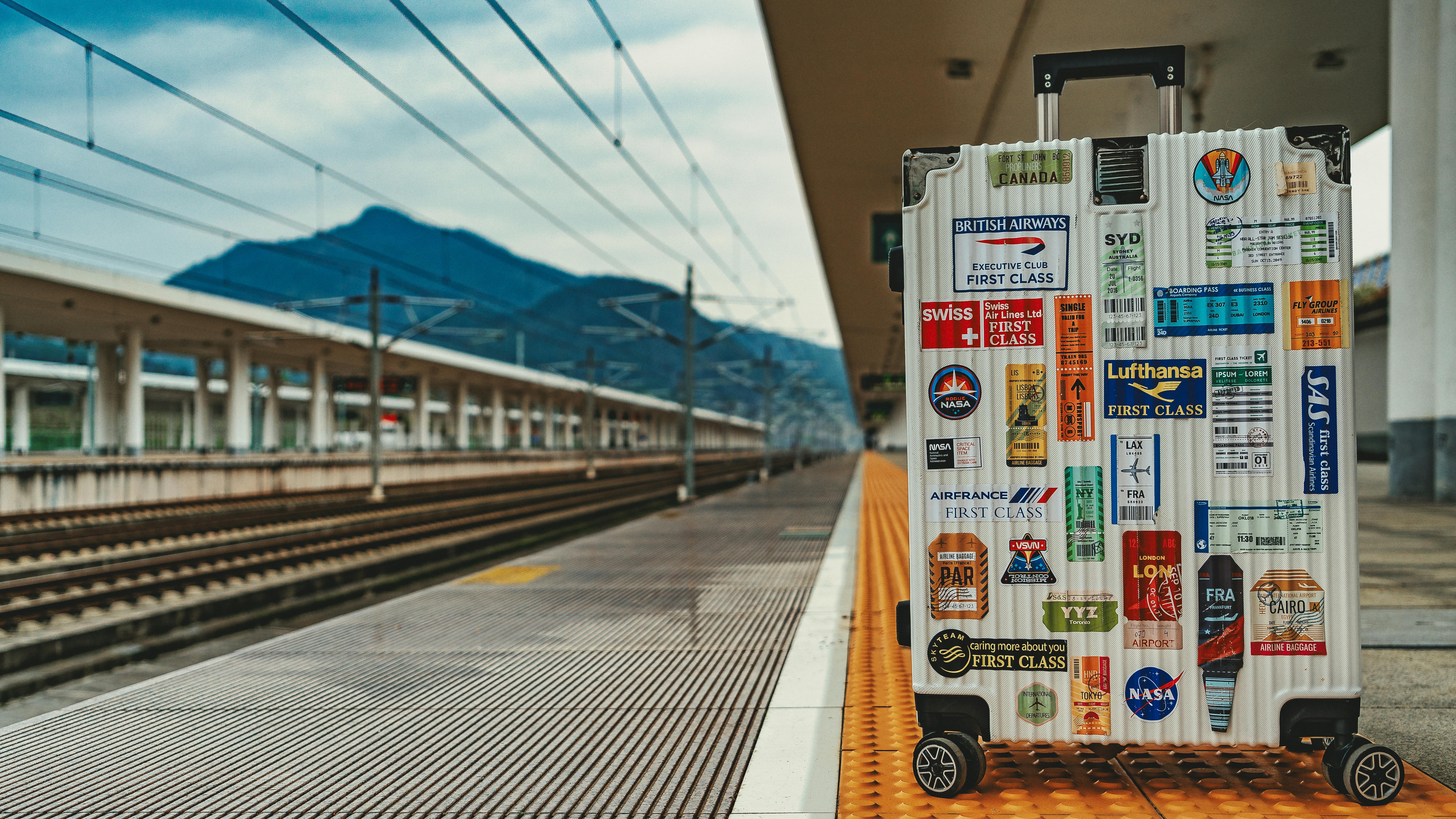 White suitcase adorned with travel stickers on empty railway platform, mountain backdrop.