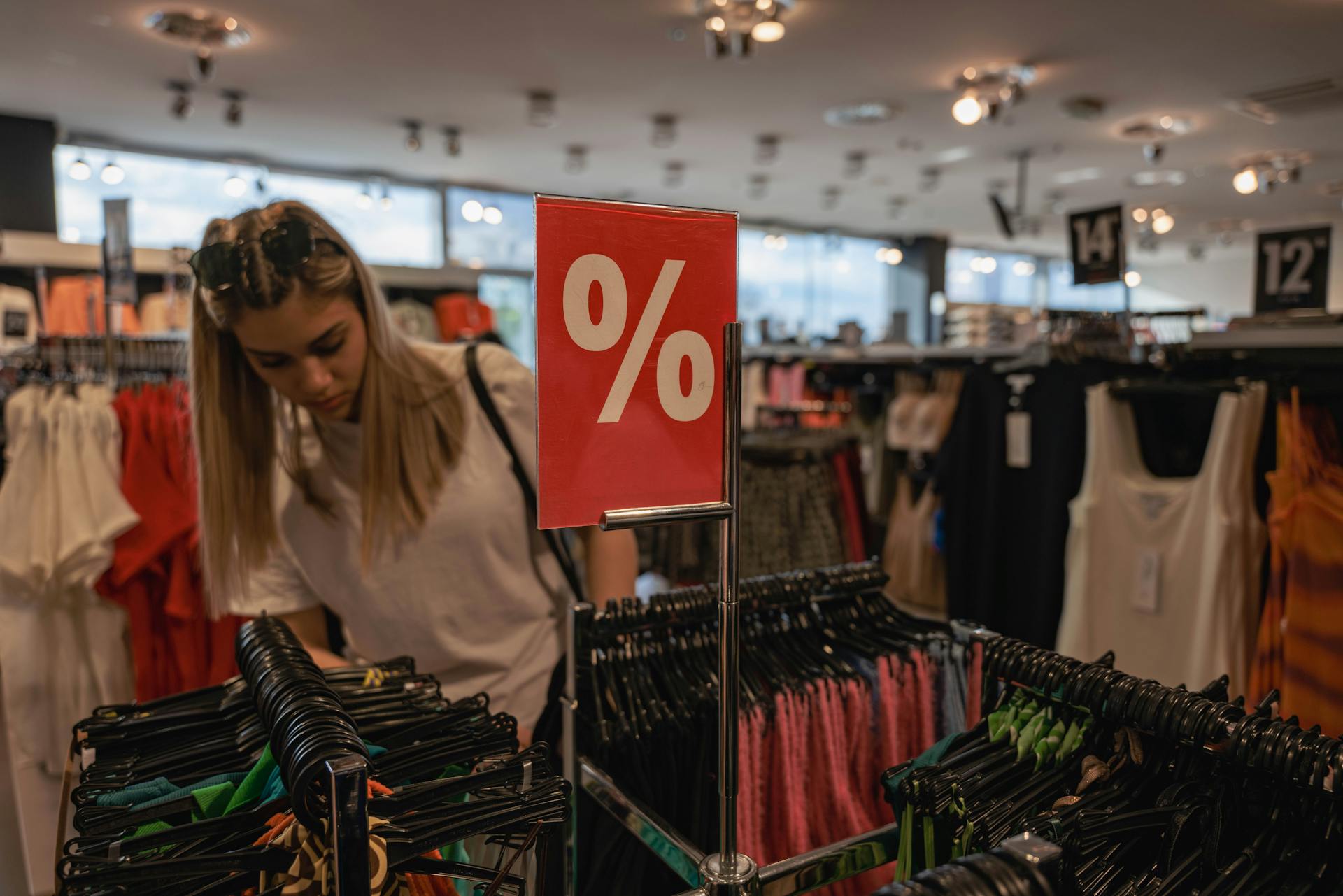 A young woman browses a clothing store during a sale, indicated by a red discount sign.