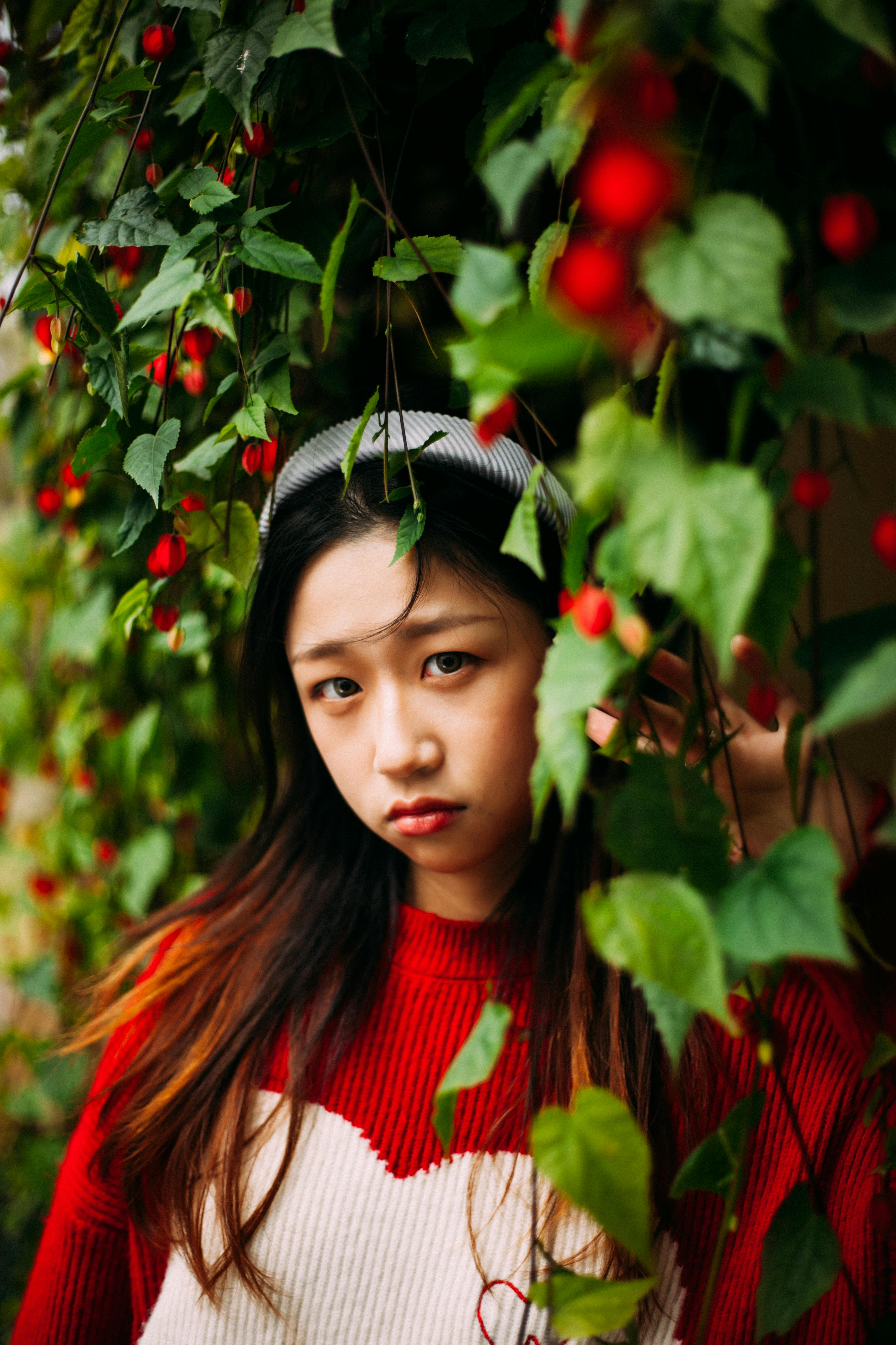 Portrait of a young woman in a red sweater surrounded by green leaves and red berries, creating a colorful and vibrant scene.