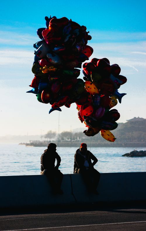 Free Two people sit by the sea holding colorful balloons against a bright sky, creating a dramatic silhouette. Stock Photo