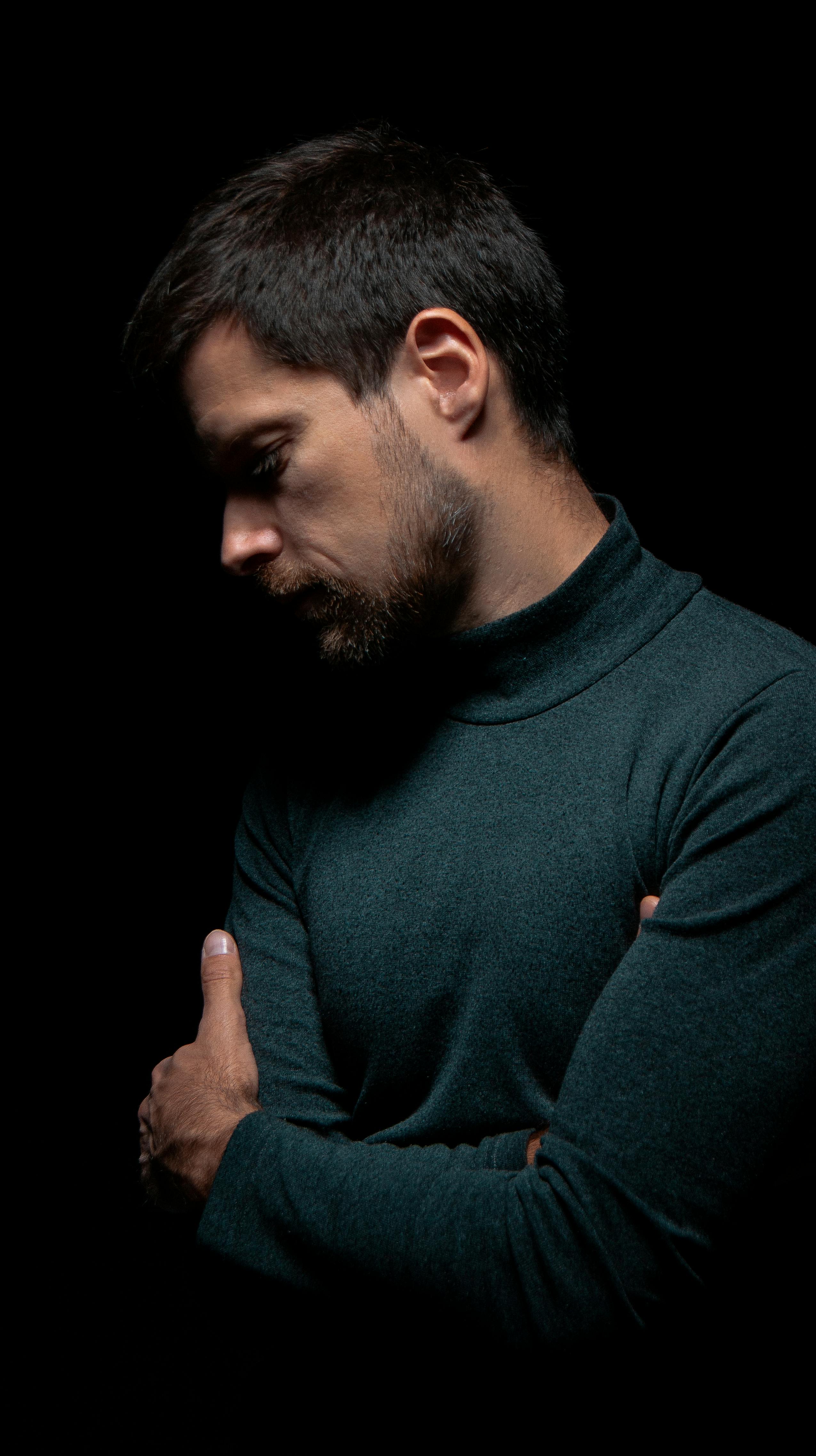 A serious man in a green turtleneck with beard and mustache poses in a dark studio setting.