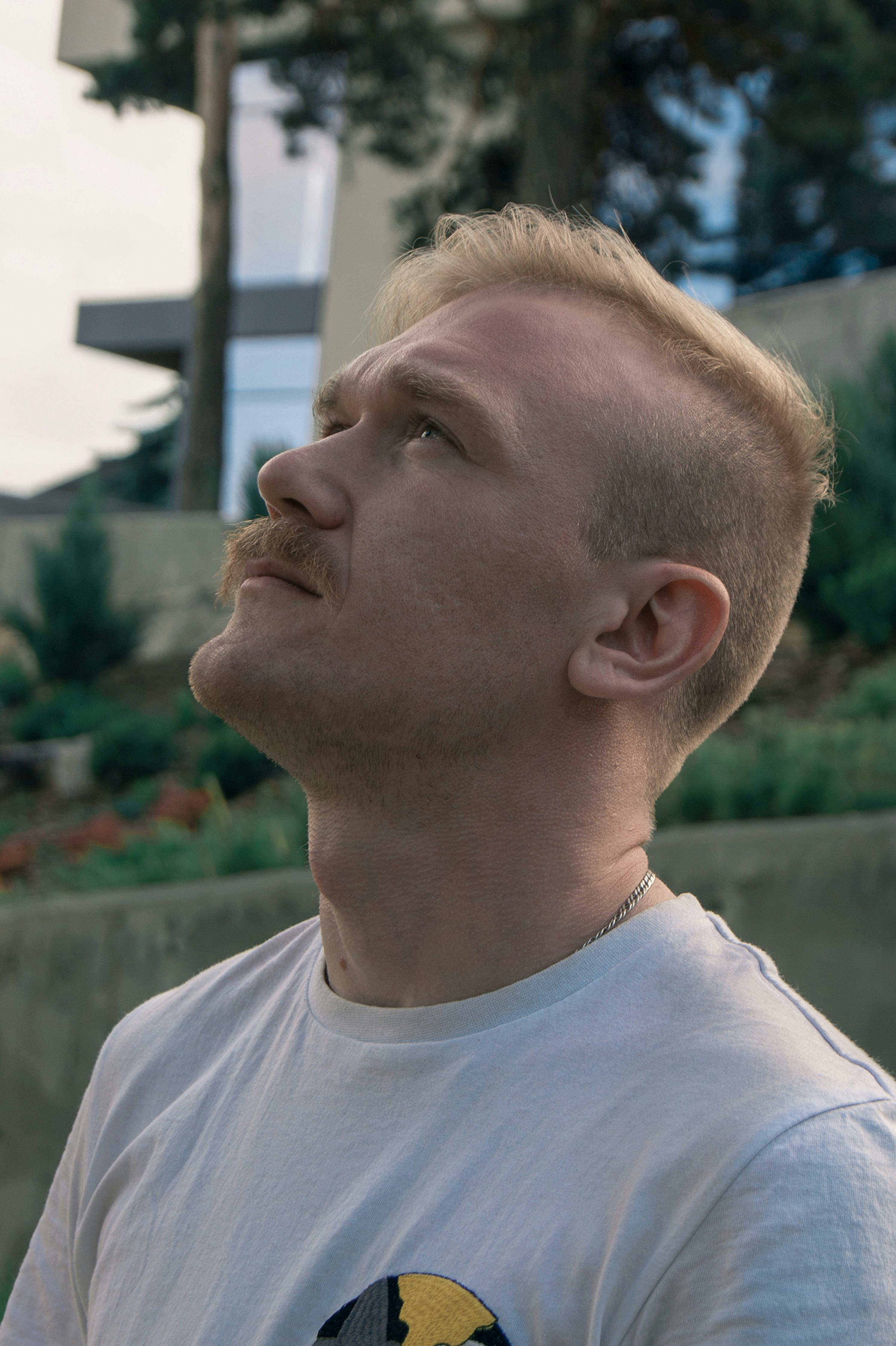 Side profile of a thoughtful man with blonde hair and a mustache, looking up outdoors.
