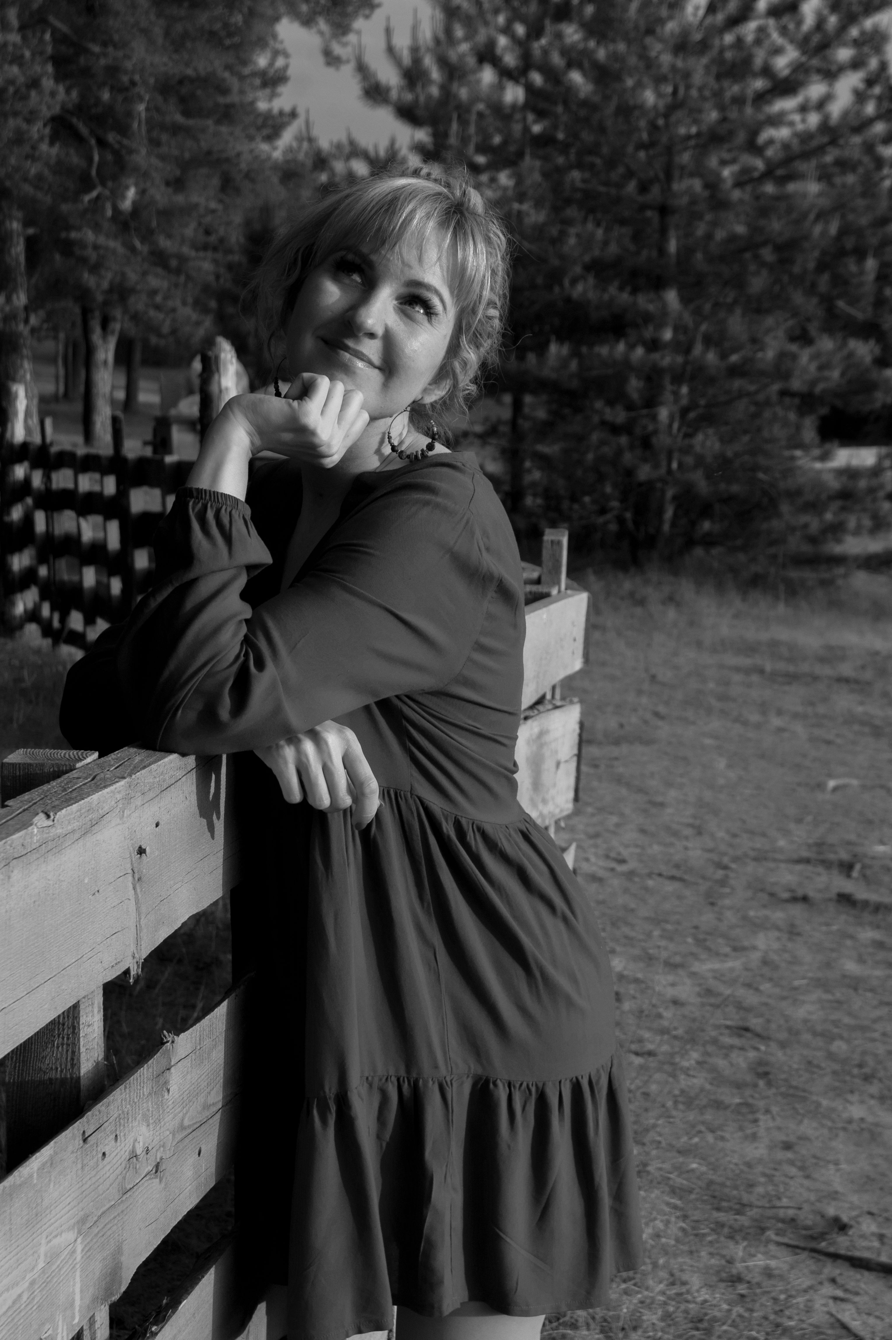 Elegant black and white portrait of woman posing stylishly against a rustic fence in a rural setting.