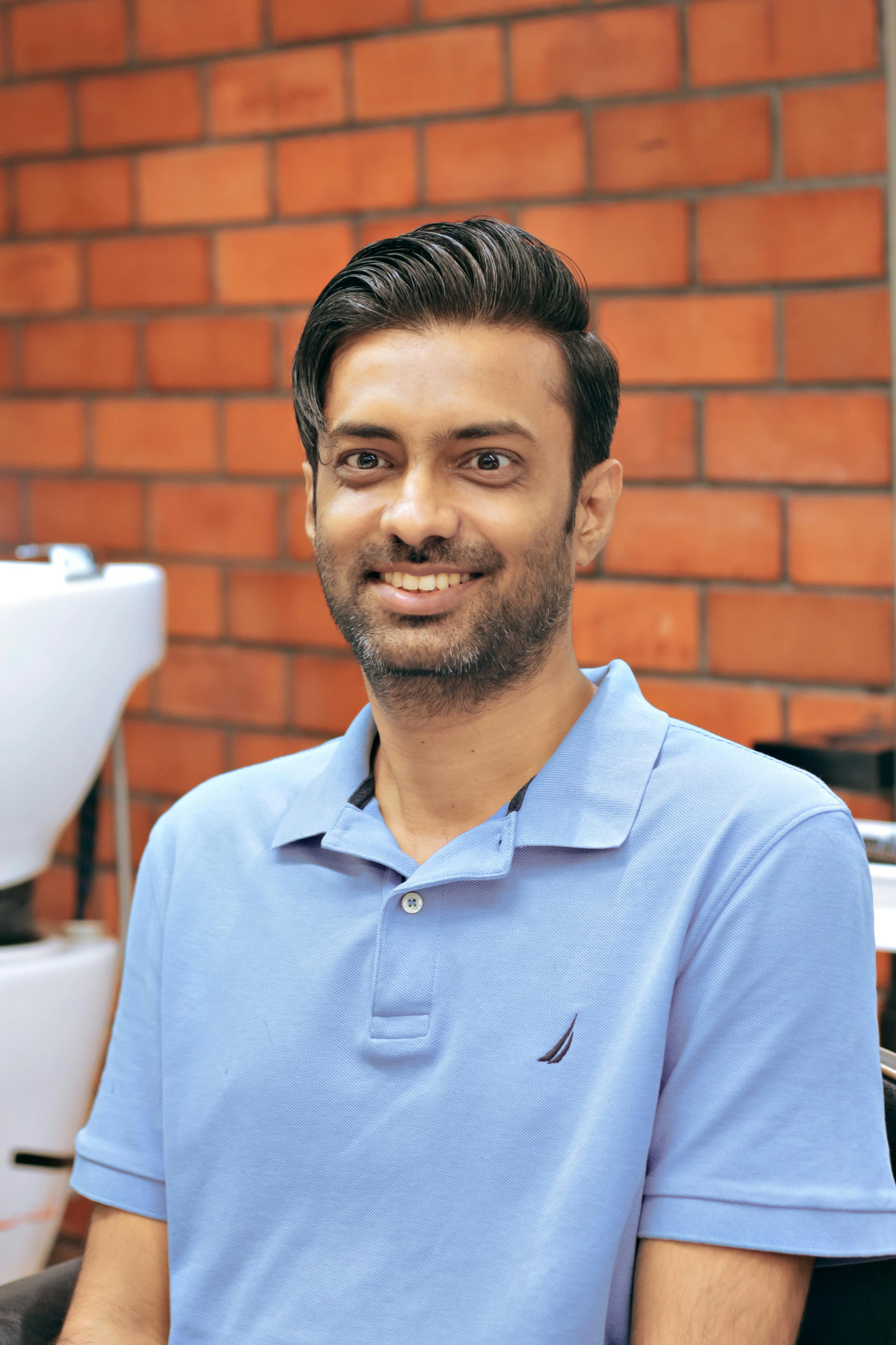 A cheerful man in a blue polo shirt sitting indoors against a brick wall background.