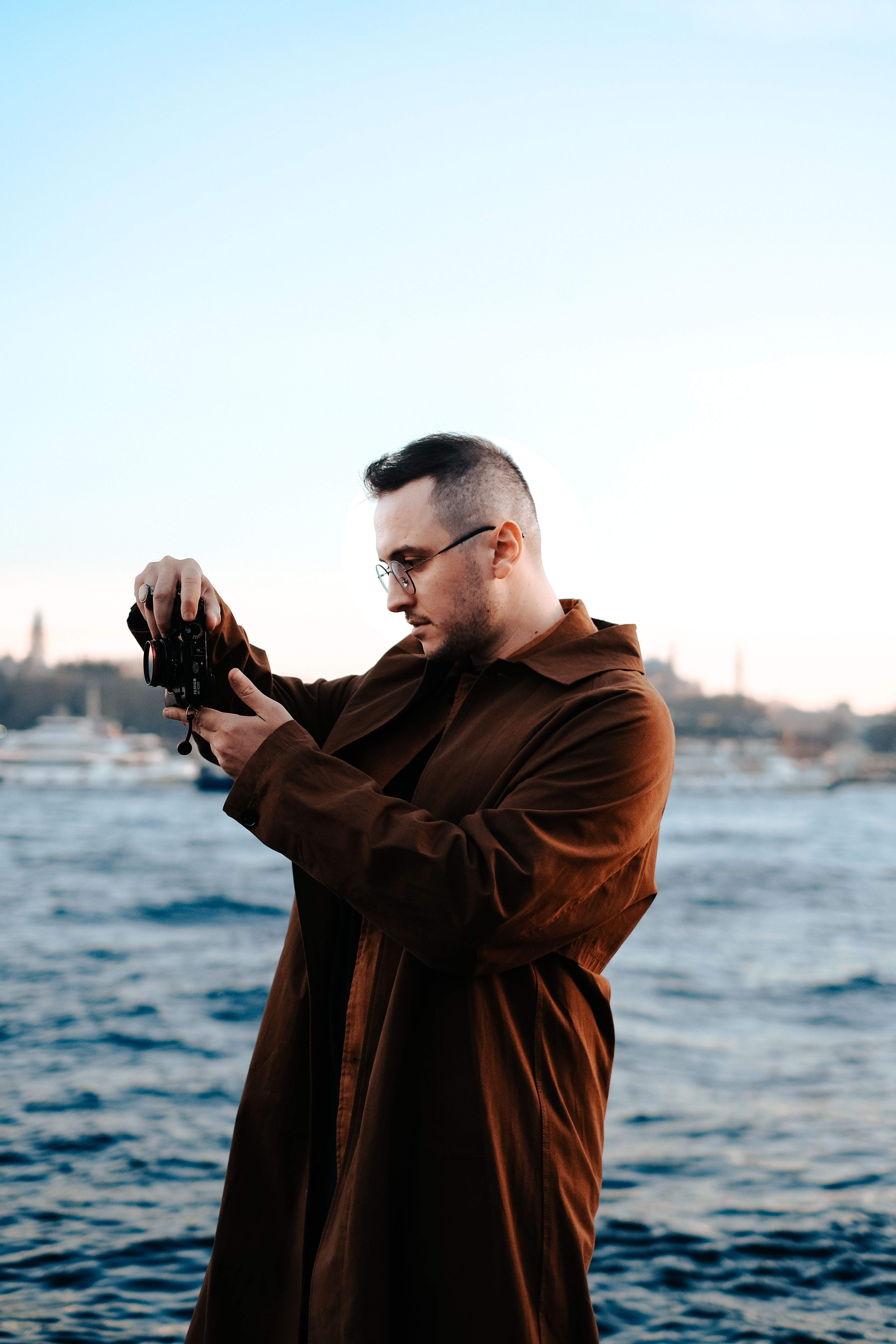 A young man in a brown overcoat captures the coastline with a camera.