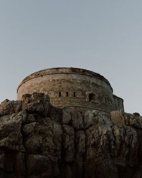 A solitary historic stone fortress stands atop a rugged rocky hill under a clear sky.