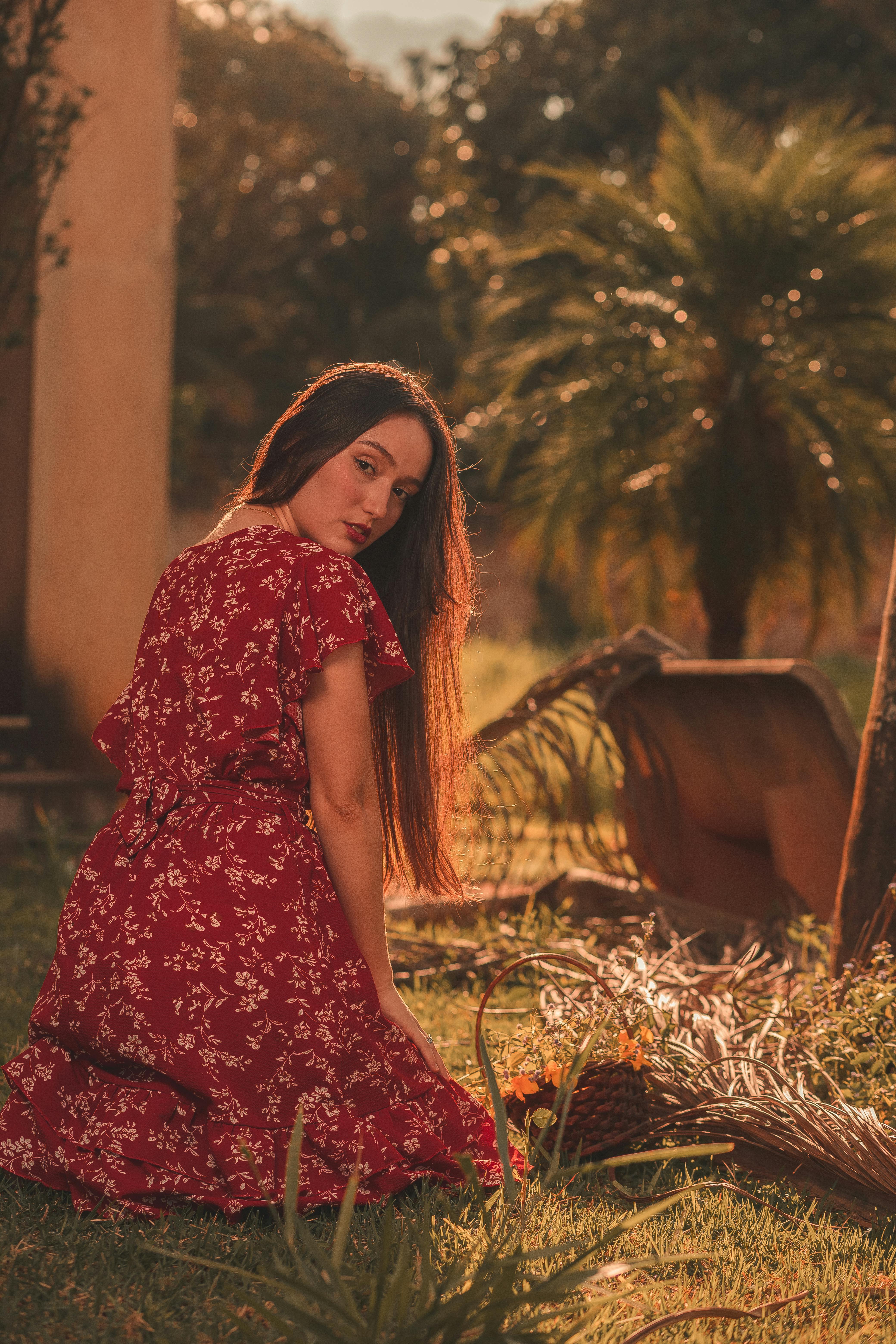 Free Woman in a stylish red dress with floral patterns sitting on grass, with a warm sunlight filter. Stock Photo