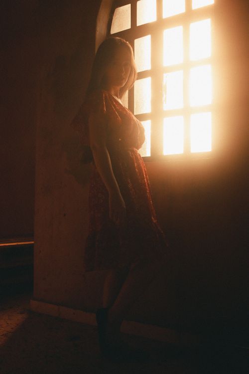 Free A mysterious silhouette of a woman in a red dress, illuminated by warm sunlight through a window. Stock Photo