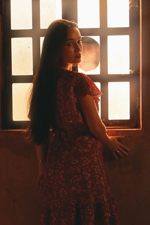 Free A woman in a floral dress poses by a softly lit window, creating a warm silhouette. Stock Photo