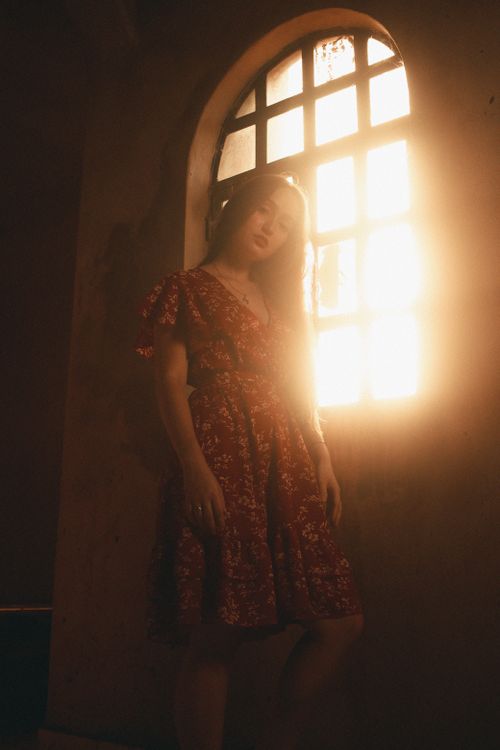 Free A woman in a red floral dress stands elegantly by a sunlit window with a rustic ambiance. Stock Photo