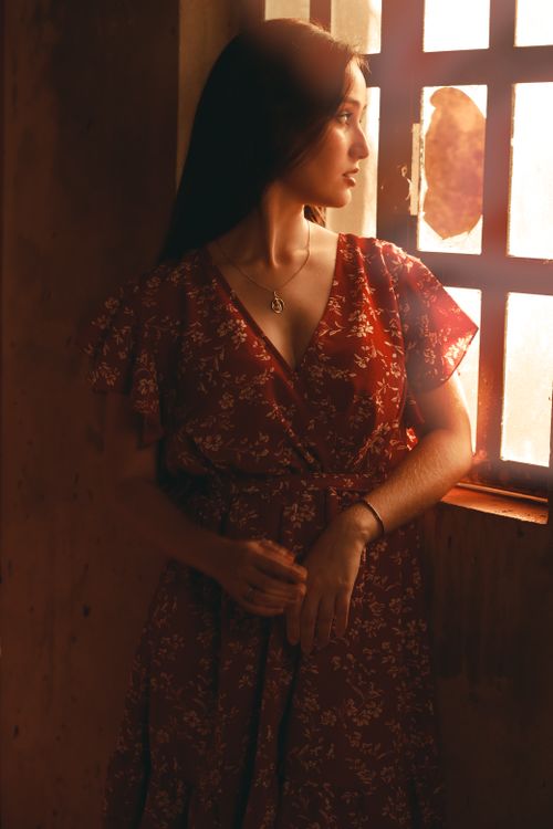 Free A woman in a red dress stands by a window, bathed in warm sunlight, creating a serene and intimate ambiance. Stock Photo