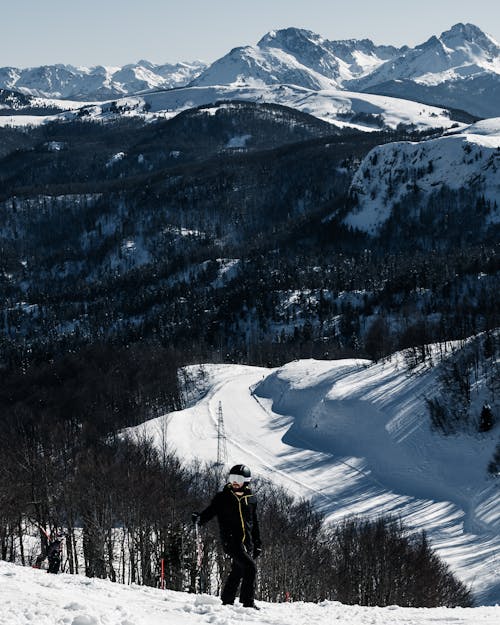 A Person in Warm Clothing and Ski Goggles Standing on a Steep Slope in Mountains 