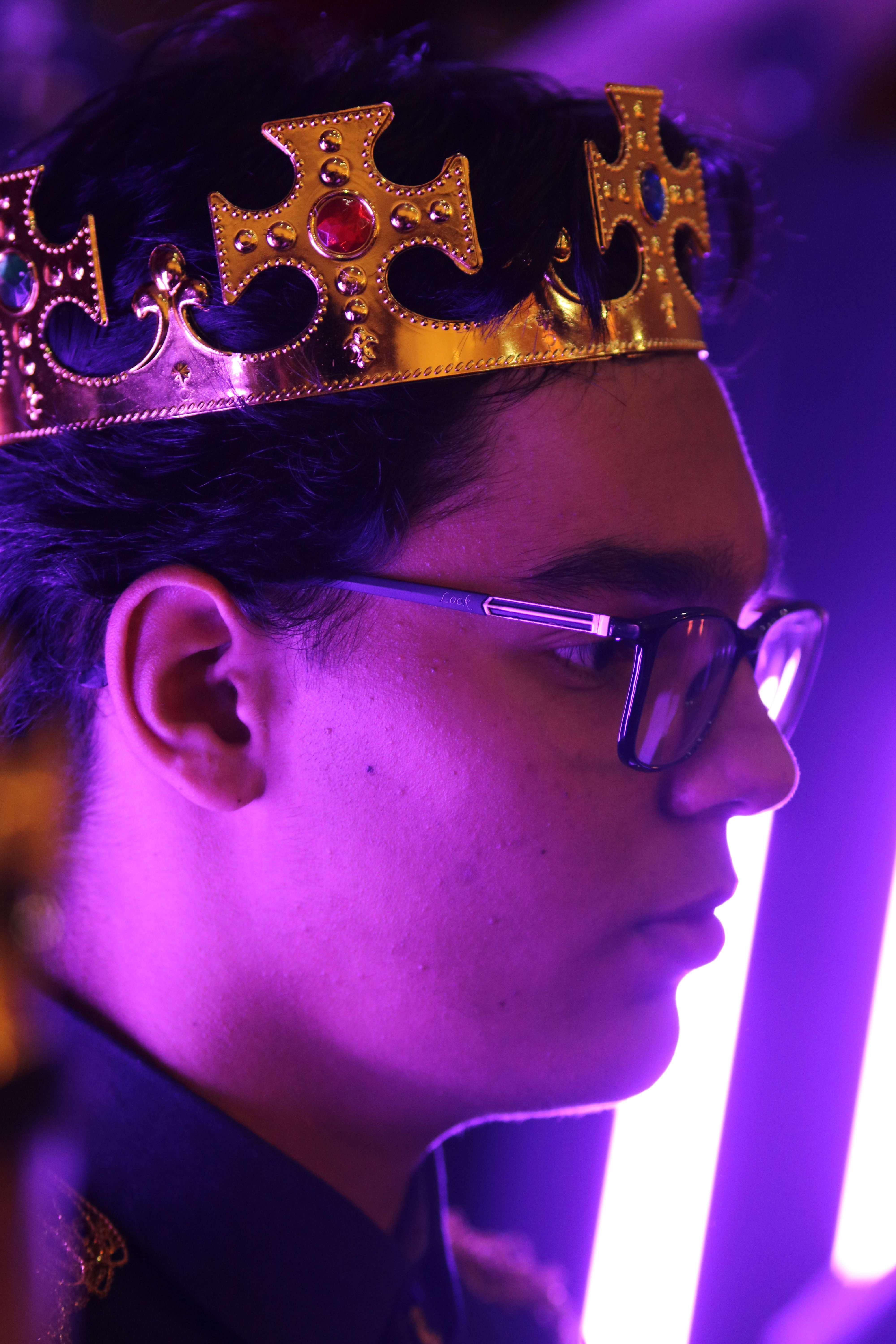 A man wearing a gold crown and eyeglasses, illuminated by purple light in a close-up portrait.