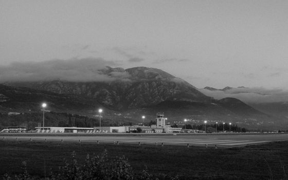 Black and white photo of Tivat Airport with illuminated runway and scenic mountains in the background.