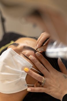 A woman with closed eyes wearing a mask receives a beauty treatment for her eyebrows.