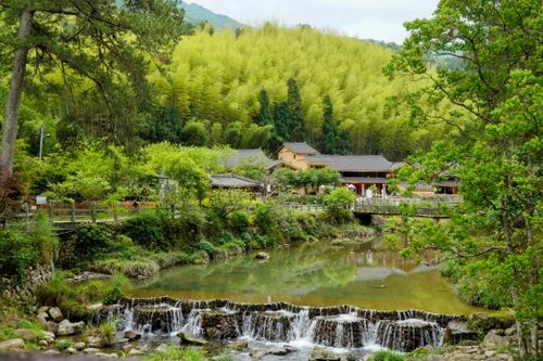 Free A serene village landscape featuring a small waterfall, lush forest, and traditional buildings under a clear sky. Stock Photo