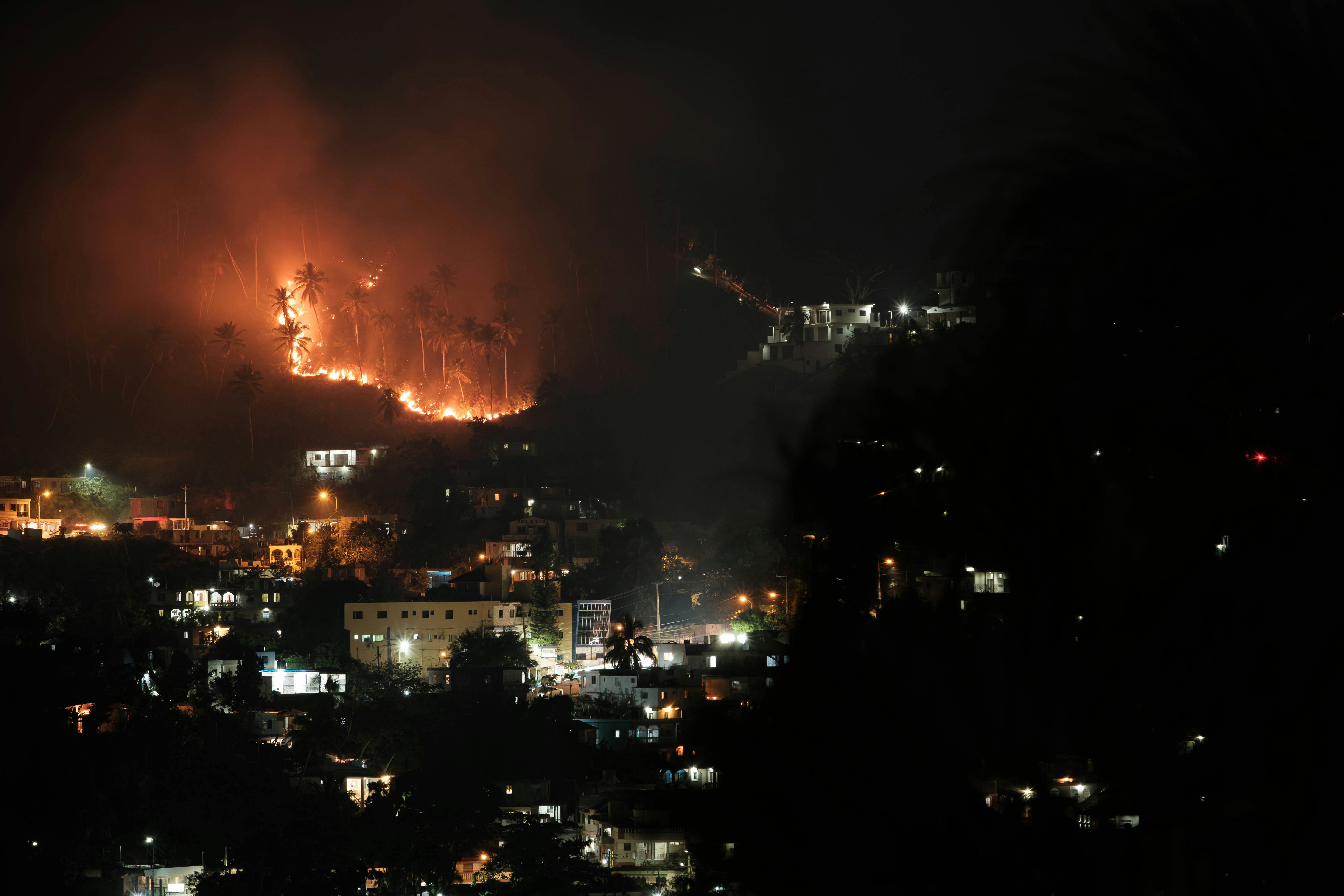 Aerial view of a forest fire at night near a town, with bright flames lighting up the sky and smoke billowing upwards.