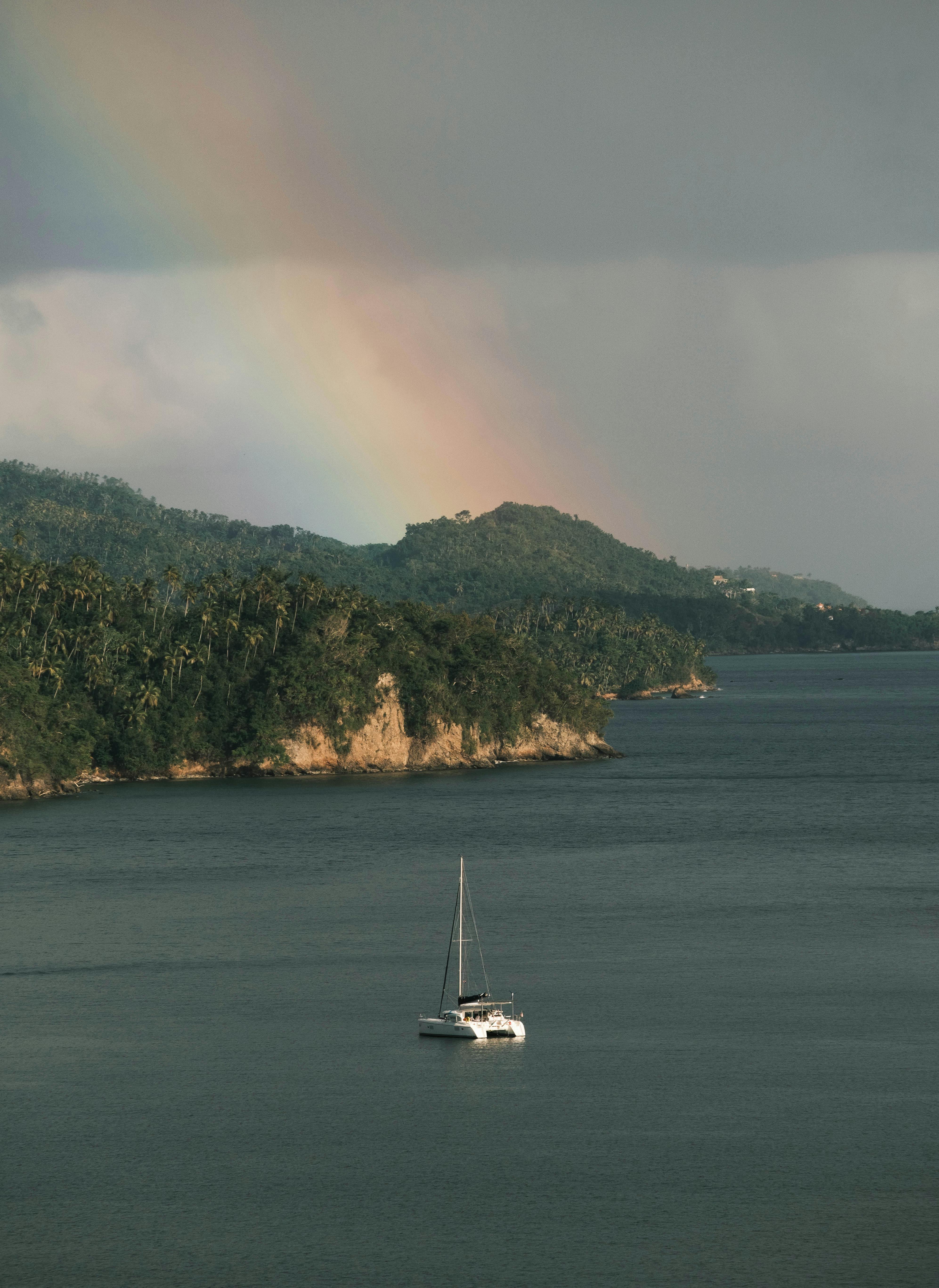 A peaceful sailboat on the ocean beneath a vibrant rainbow arching over lush coastal hills.