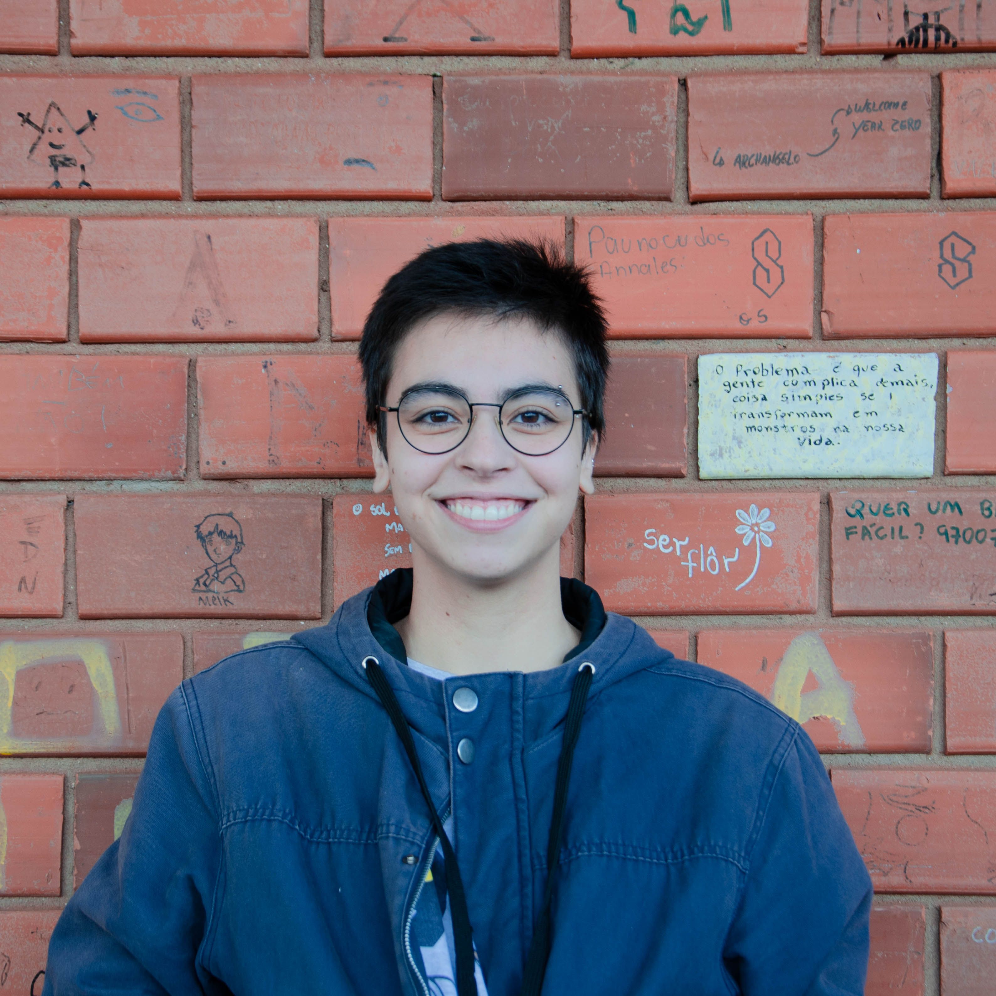 Portrait of a smiling person with glasses and short hair in front of a colorful brick wall with graffiti.