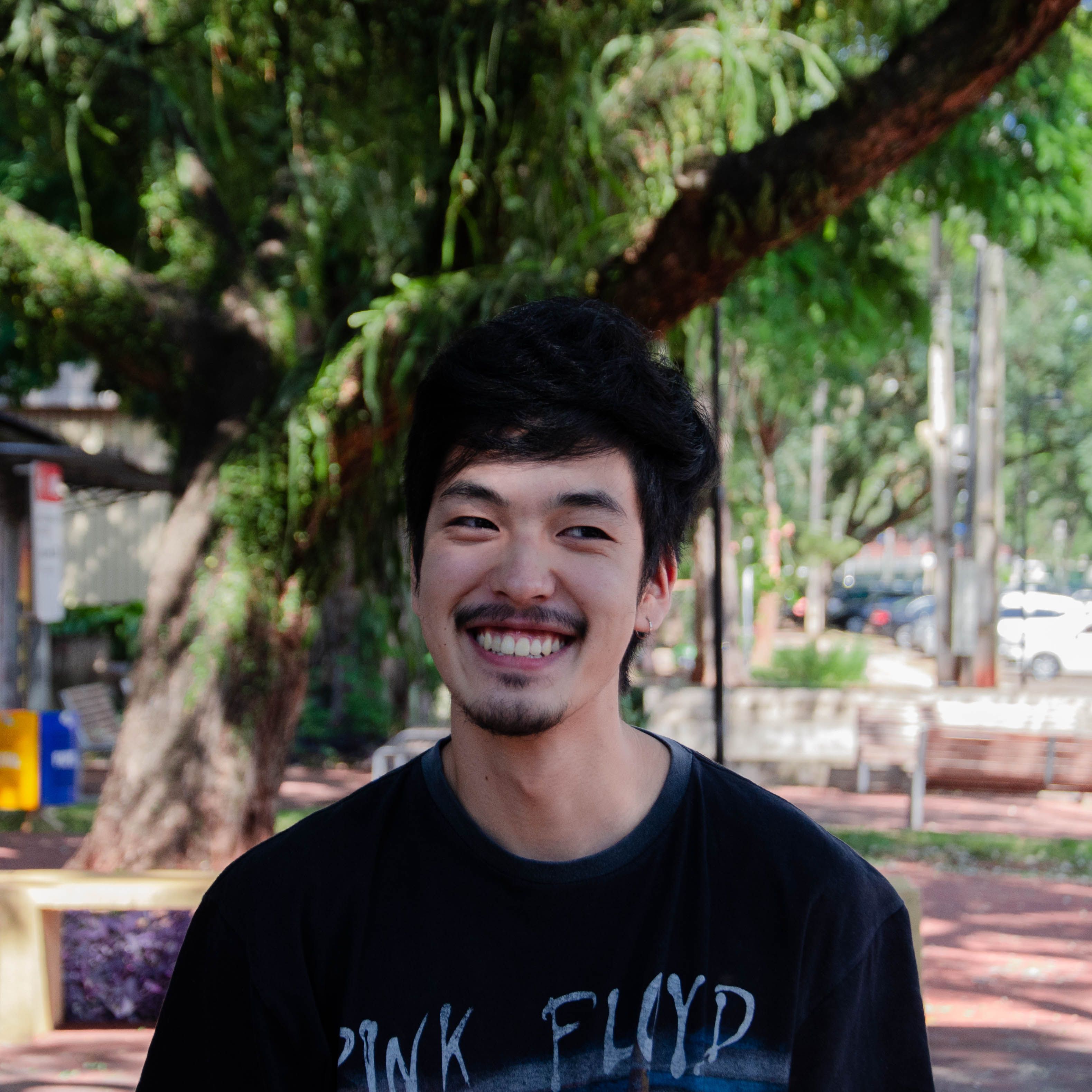 A cheerful young man with a beard smiling in a sunny urban park, wearing a Pink Floyd t-shirt.