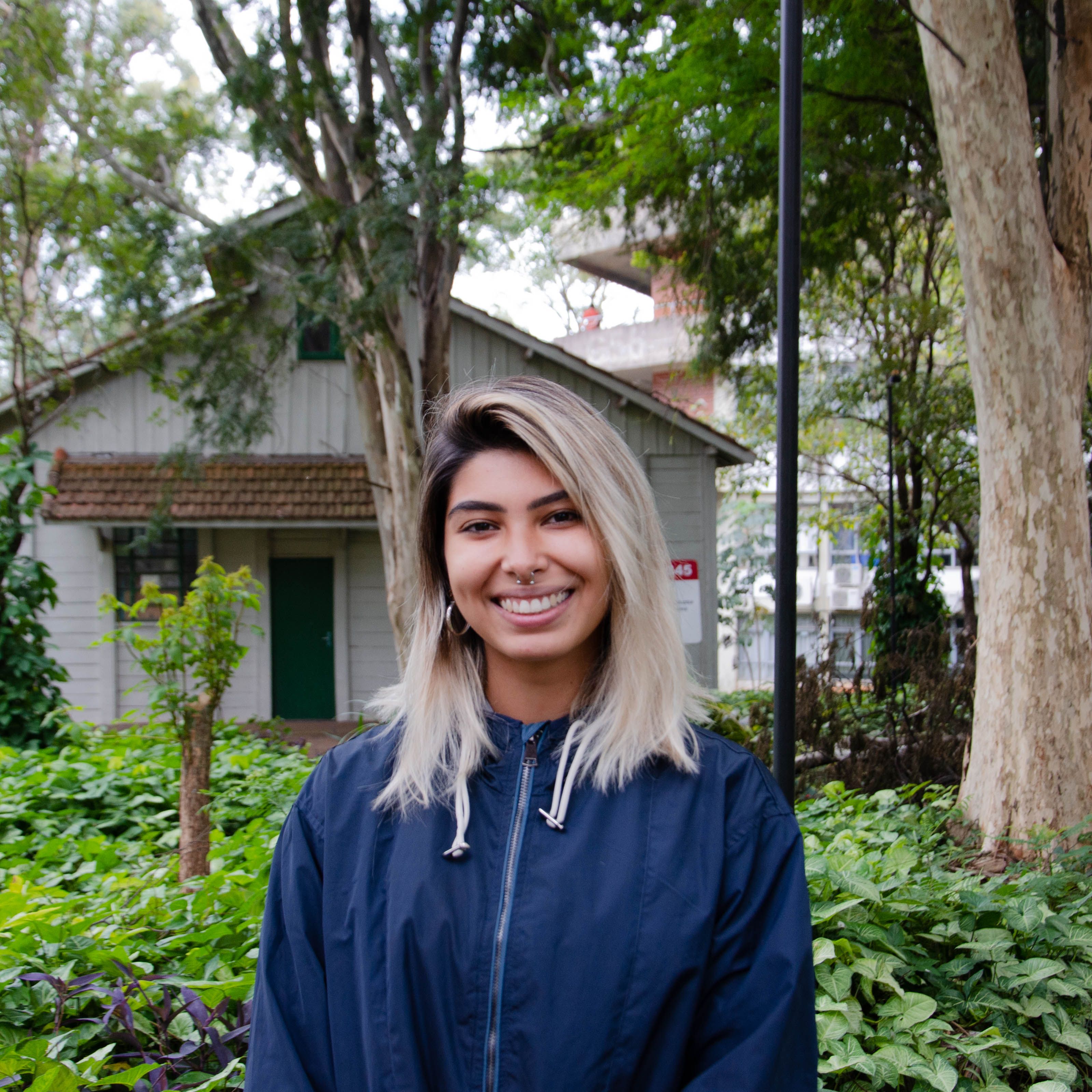 A woman with blonde hair smiles at the camera, standing in a lush green garden with trees and a rustic house in the background.