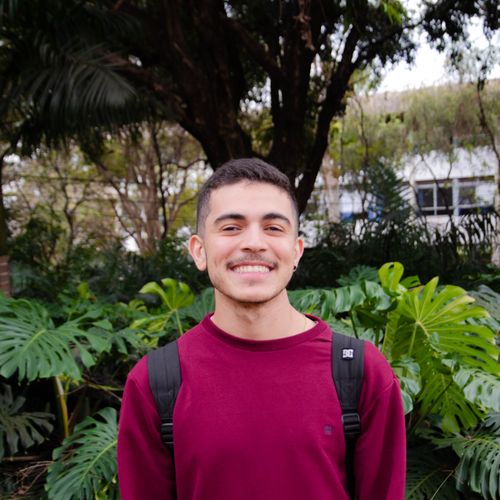 Free Cheerful young man wearing a backpack, standing in front of lush foliage outdoors. Stock Photo
