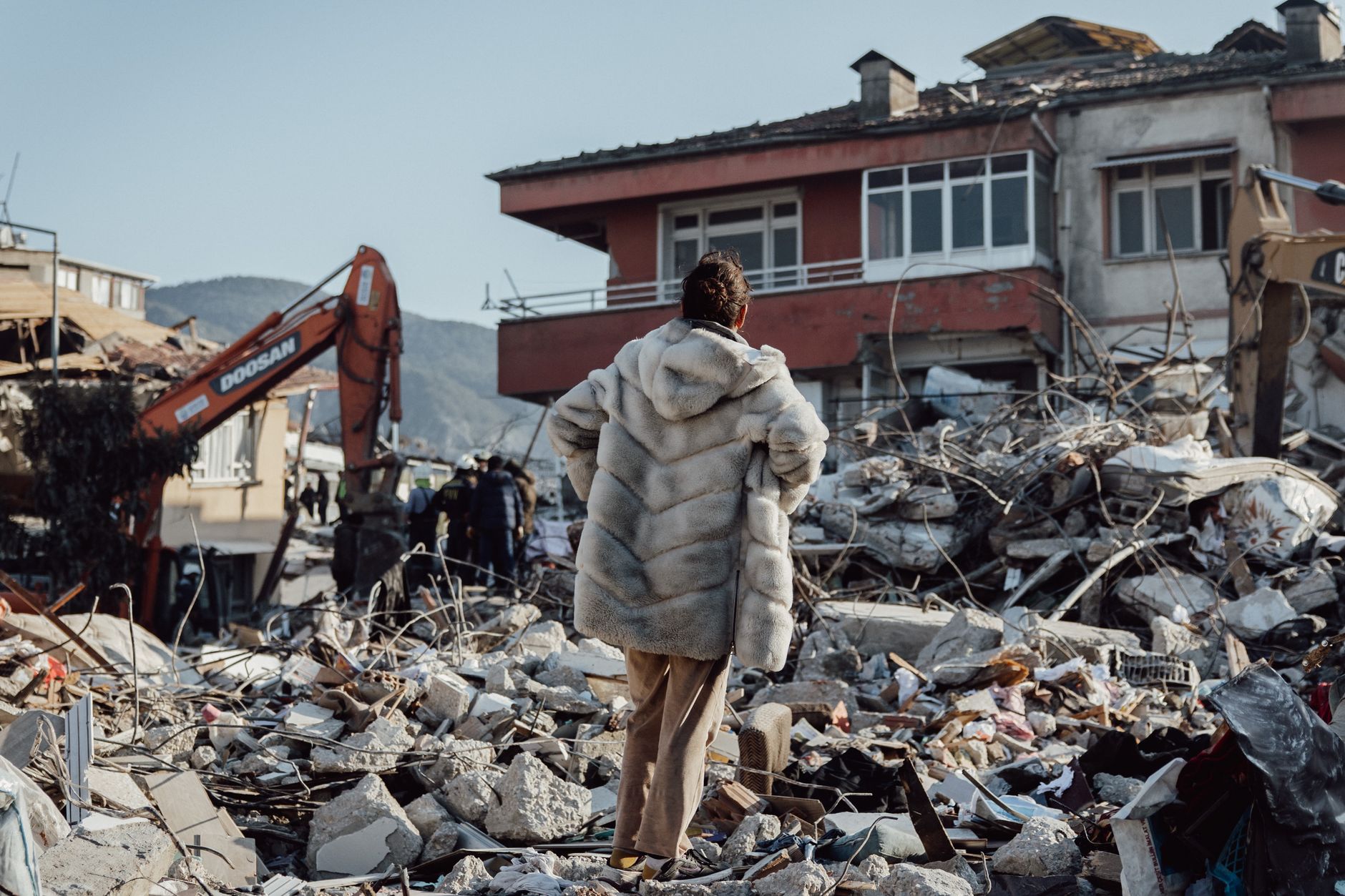 A woman in a fur coat stands amidst earthquake rubble, observing damaged buildings and an excavator.