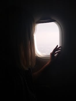 A woman silhouetted against the bright sky, gazing through an airplane window during flight.