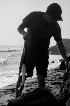 A black and white silhouette of a fisherman holding a catch on a Mexican beach.