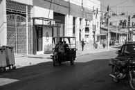 Black and white street view in Pinar del Río, featuring a rikshaw and pedestrians on a sunny day.