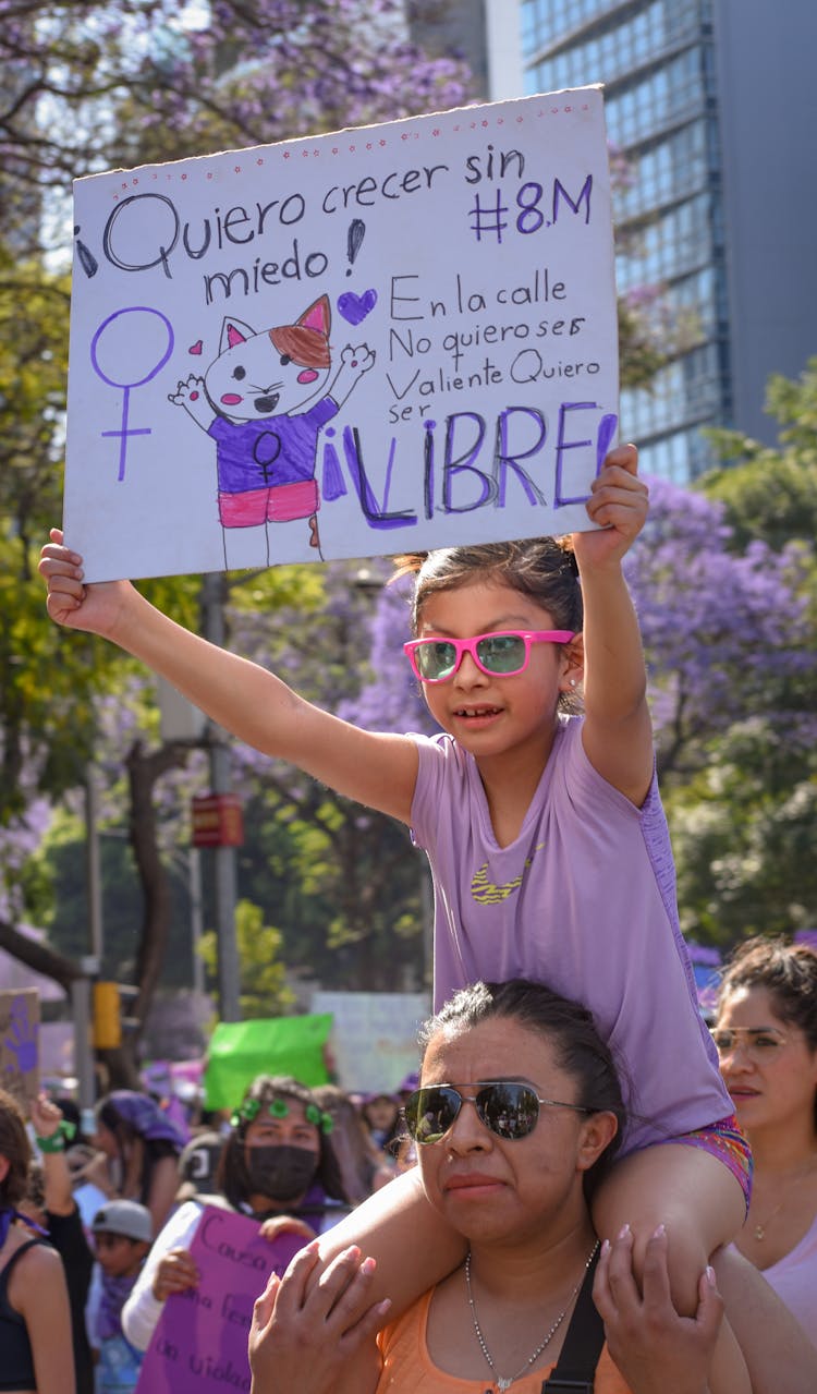 Little Girl With A Banner Sitting On Her Mothers Shoulders At A Rally On Womens Day