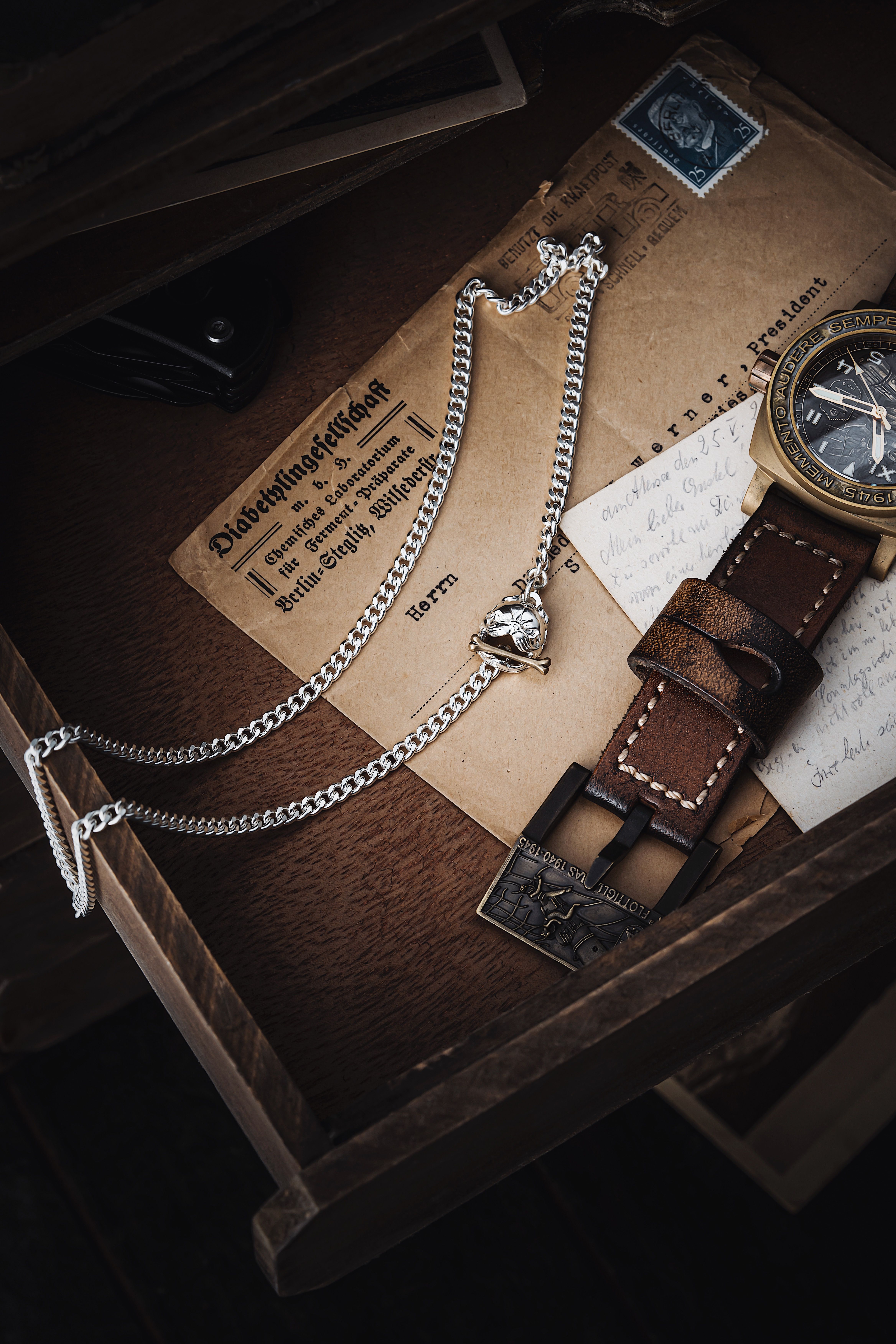A vintage watch and silver necklace in a wooden drawer with old letters and stamps.
