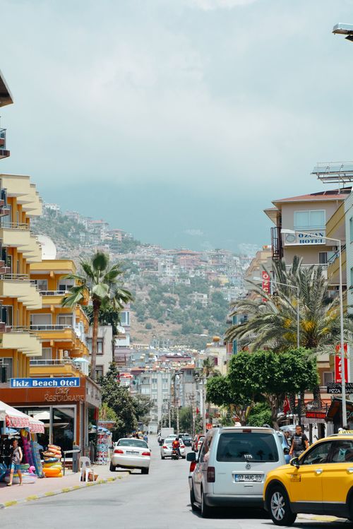 Free Bustling street scene in Alanya, Türkiye with hotels, cars, and a mountainous backdrop. Stock Photo