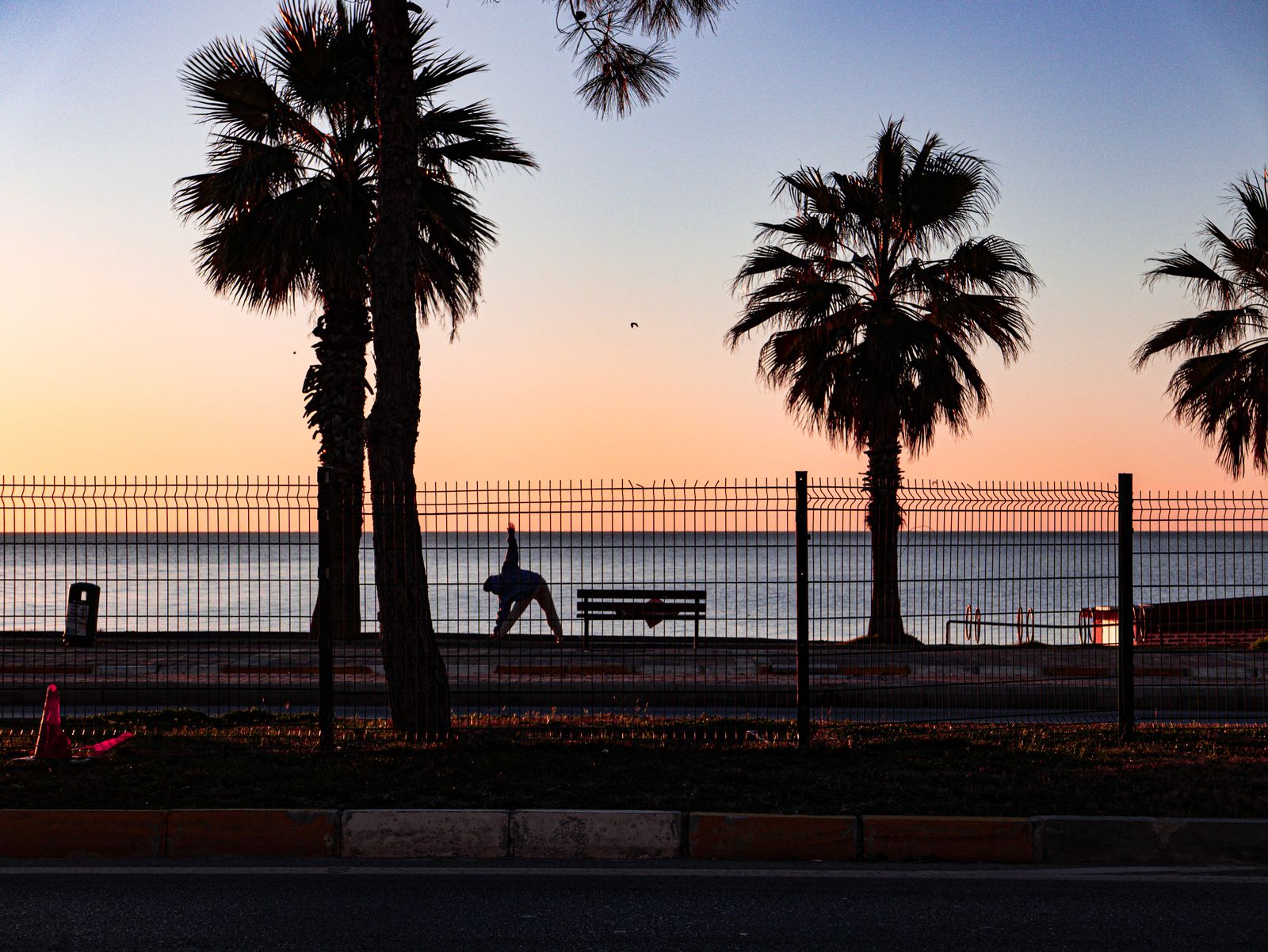 Silhouette of a person practicing a yoga pose by the ocean at sunset framed by palm trees
