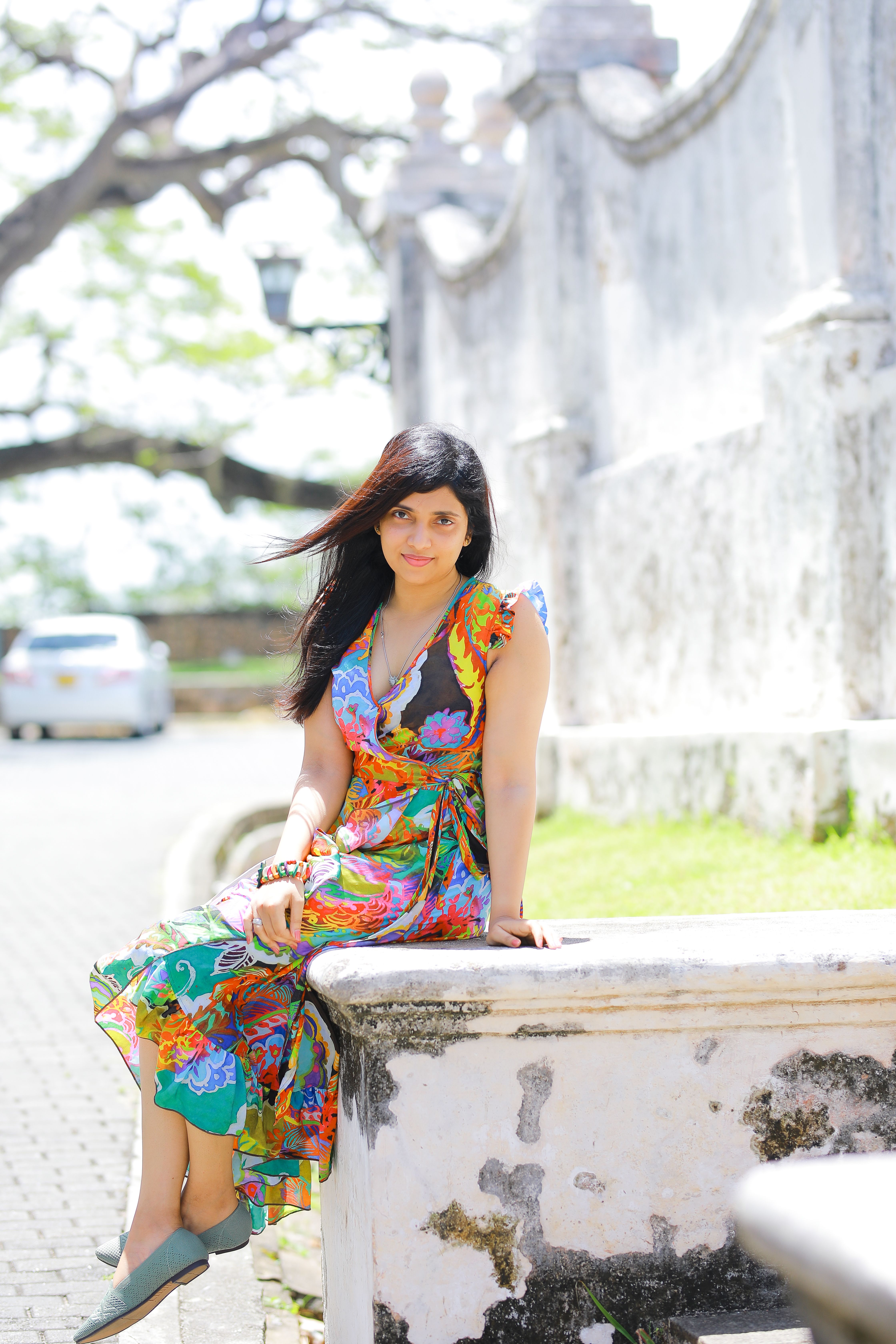 Woman in colorful dress sitting by park wall, enjoying a sunny day.