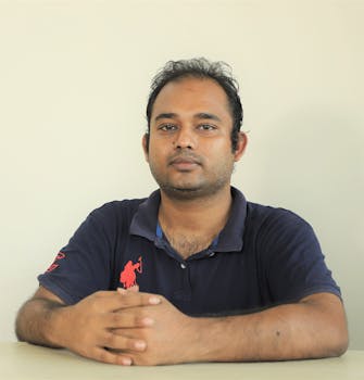 Portrait of a confident man sitting casually in a blue polo shirt against a neutral background.