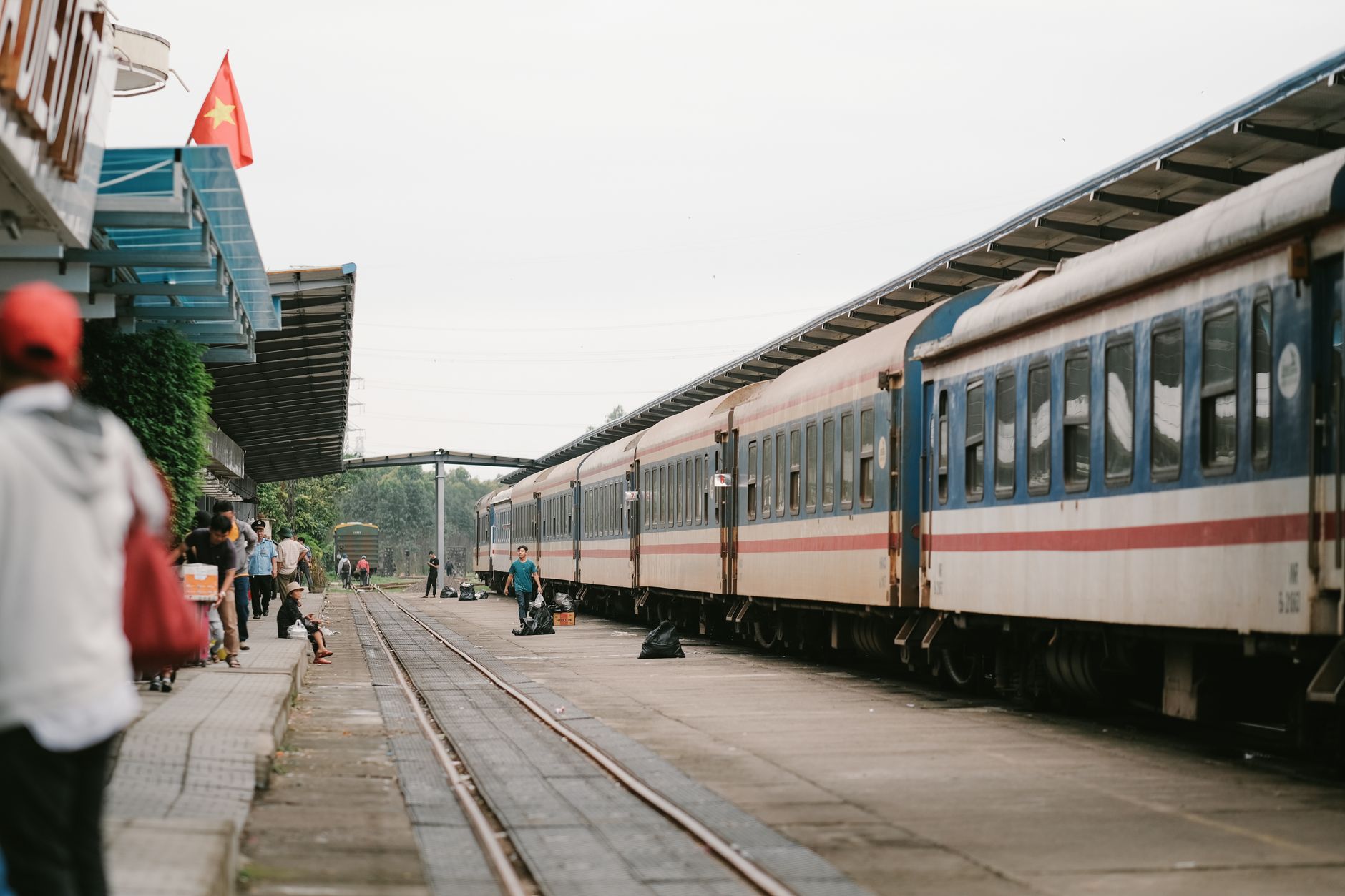A bustling railway platform with a passenger train and commuters under a cloudy sky.