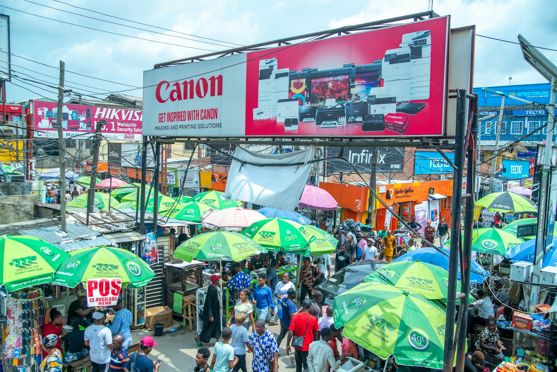 Lively market scene in Lagos with vibrant umbrellas and bustling crowds under a Canon advertisement.