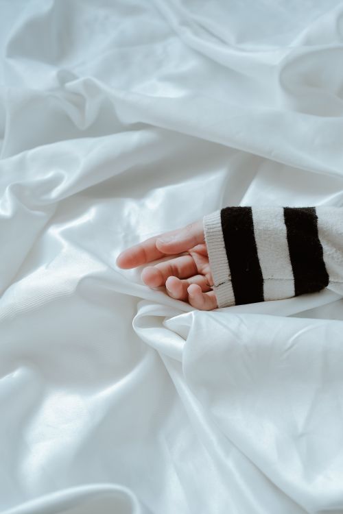 Free Close-up of a hand in a sweater resting on luxurious white satin sheets, evoking relaxation. Stock Photo