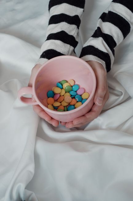 Hands holding a pink mug filled with colorful candies on a soft fabric background.